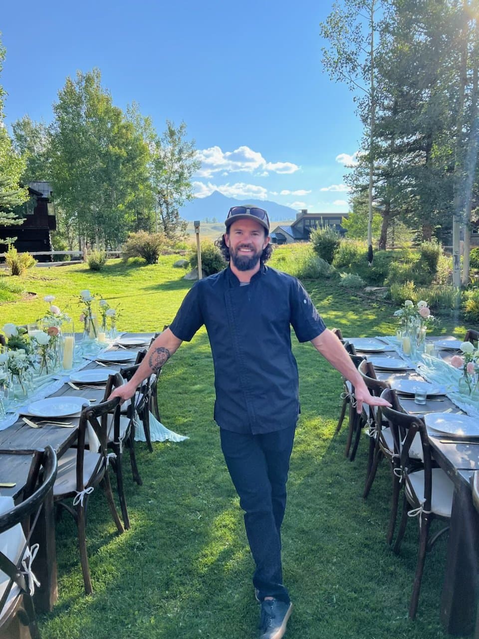 Chef Jake standing between two long outdoor catering tables set for dinner, mountains and open sky behind him