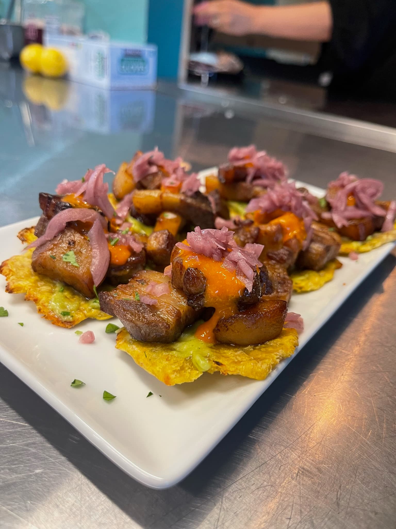 A platter of crispy plantain rounds topped with caramelized braised meat, orange sauce, and pickled red onions