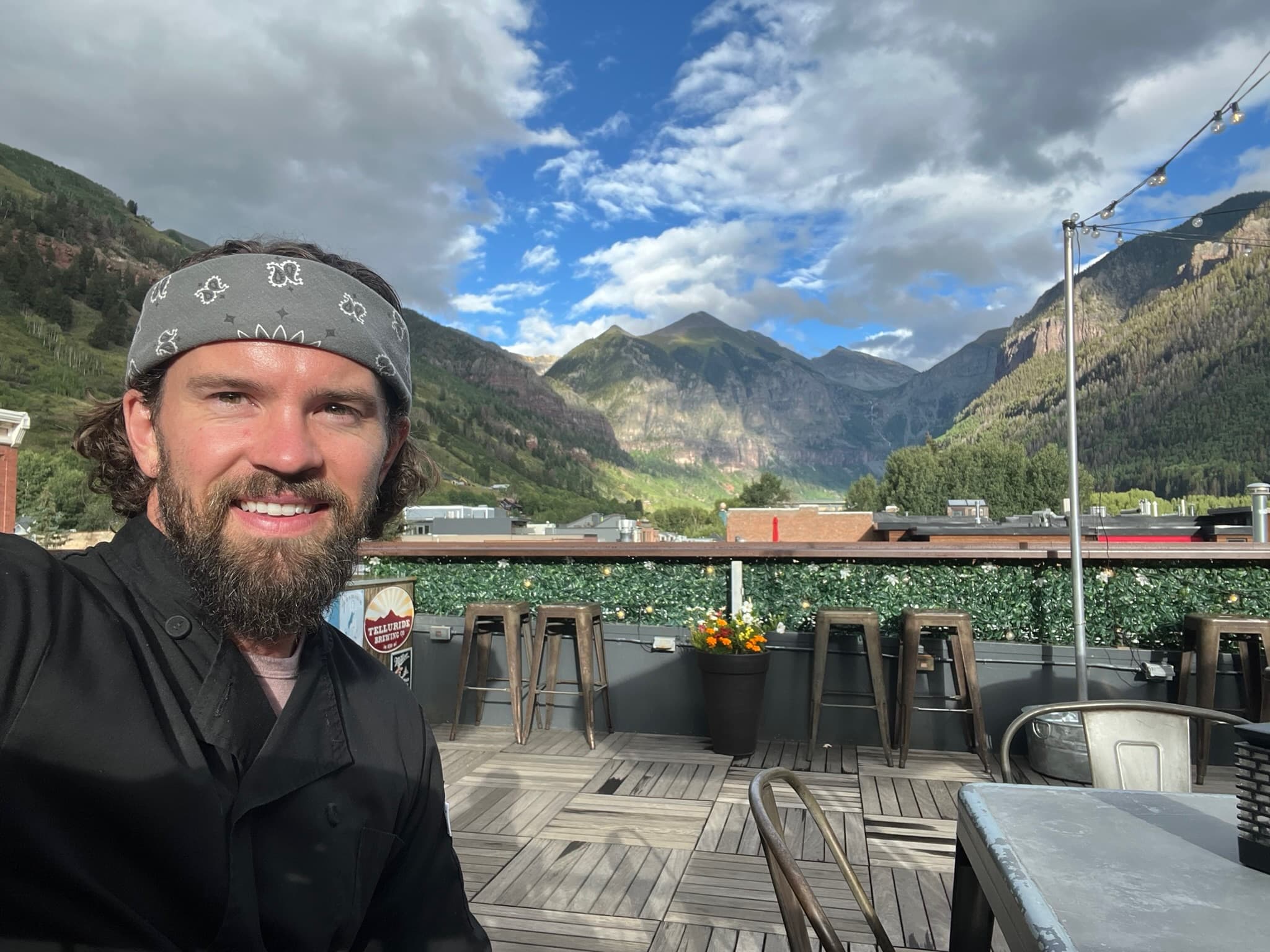 A bearded man wearing a gray bandana smiles at a rooftop restaurant with mountains and forested valley visible in the background.
